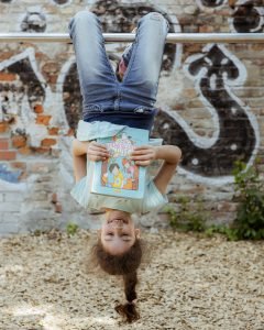 Ein Mädchen mit langem Zopf, kopfüber mit den Knien an einer Stange hängend, liest dabei ein Buch.