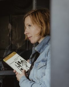 Eine ca. 40-jährige Frau mit hellen, kinnlangen Haaren und Jeansjacke. Sie hält ein geschlossenes Buch in der Hand, der Blick ist entspannt und offen.