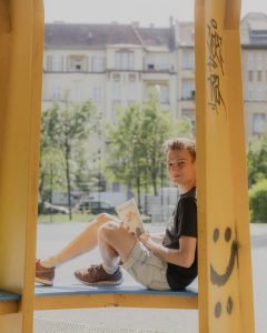 Ein junger Mann in T-Shirt und kurzer Hose sitzt auf einem bunten Holzgestell, hält ein Buch in der Hand und blickt offen in die Kamera