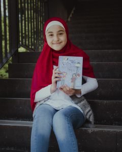 Ein freundlich lächelndes Mädchen mit rotem Hijab und blauen Jeans hält ein Buch in die Kamera