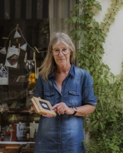 Eine Frau mit glatten langen grauen Haaren, in Jeans gekleidet, zeigt auf das Titelbild des Buches in ihrer Hand, im Hintergrund das Schaufenster einer Buchhandlung