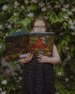 Ein Mädchen in einem Kleid steht vor einem blühenden Strauch und hält ein großes Bilderbuch in beiden Händen
